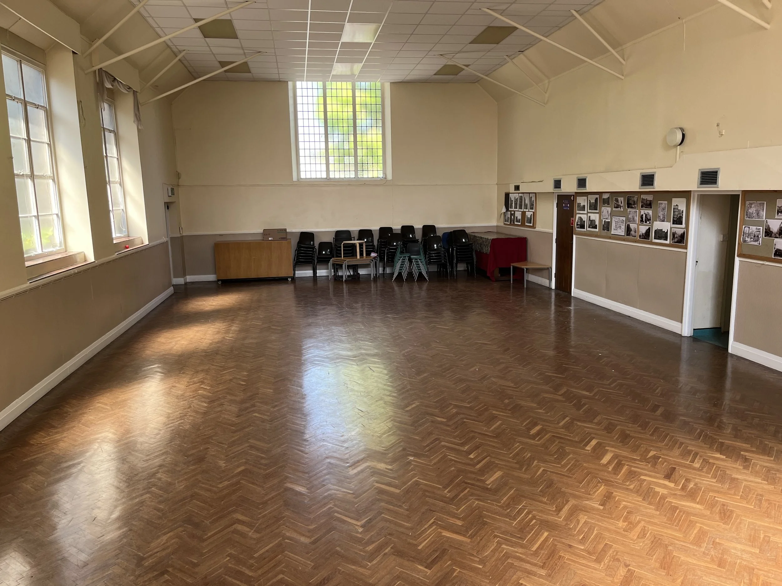 Empty room with wooden flooring, beige walls, large windows, and stacked black chairs against the wall.