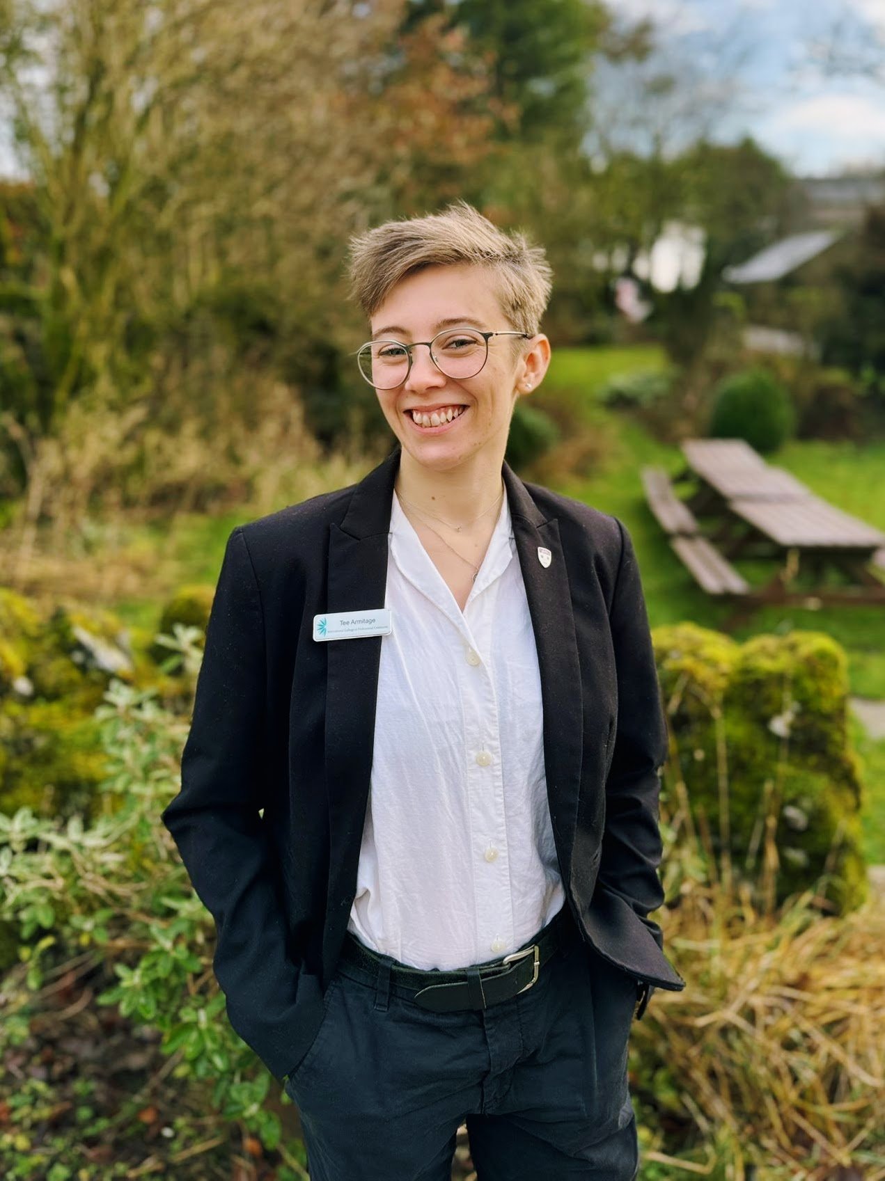 A young woman with short light brown hair and glasses, smiling outdoors in a garden with green bushes and trees, wearing a black blazer, white shirt, and black pants.