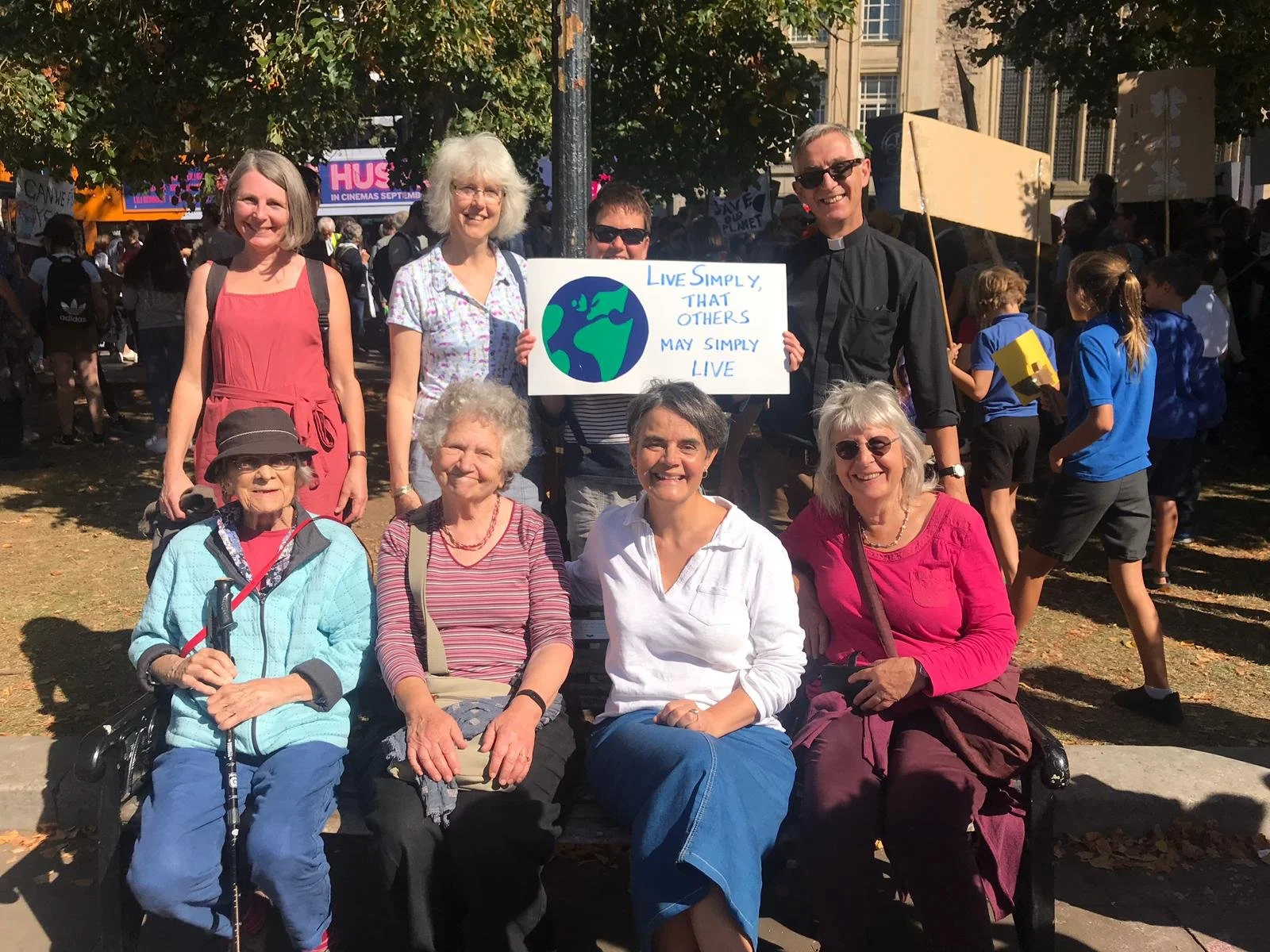 Group of people sitting and standing at an outdoor event or rally with some holding a sign that reads "Live Simply, That Others May Simply Live" and an illustration of the Earth, with a crowd of young people and trees in the background.