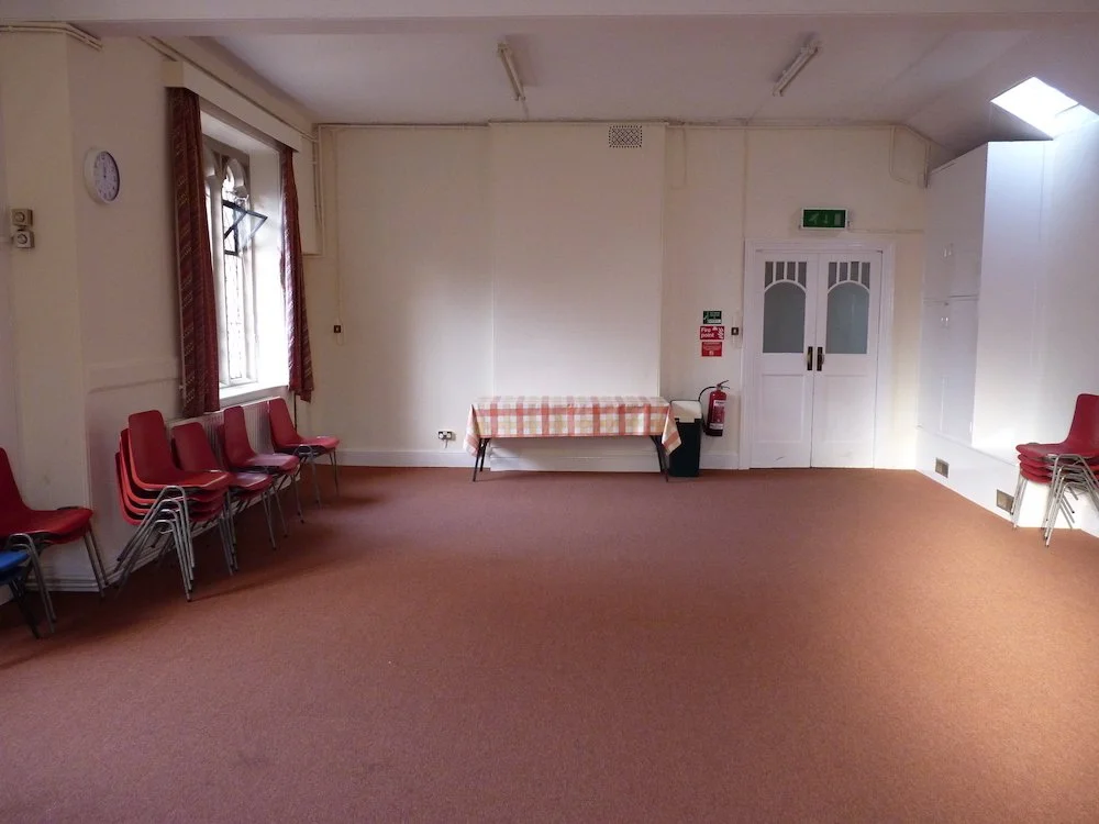 An empty room with red chairs stacked against the wall, a window with curtains, a clock, a table with a checkered tablecloth, fire safety equipment, and a door.