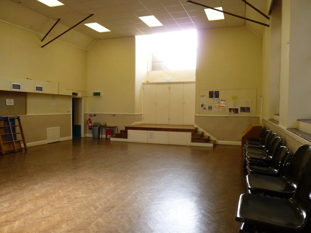 Empty room with black chairs lined up along the right wall, a small stage at the back, and bulletin boards on the walls, illuminated by natural light from a high window.