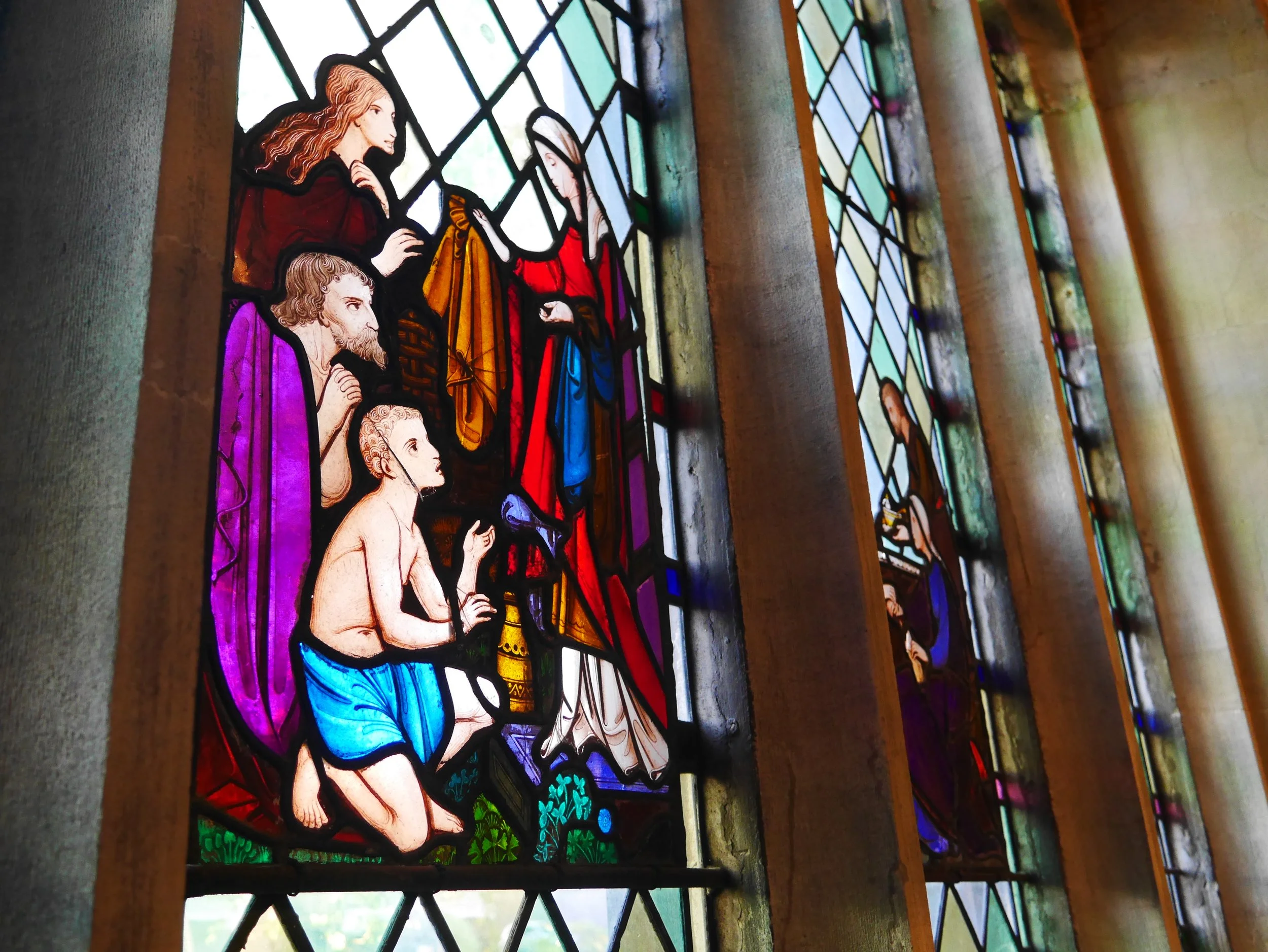 Colorful stained glass window depicting a religious scene with several figures, including a kneeling boy and others standing around him, inside a church or chapel.