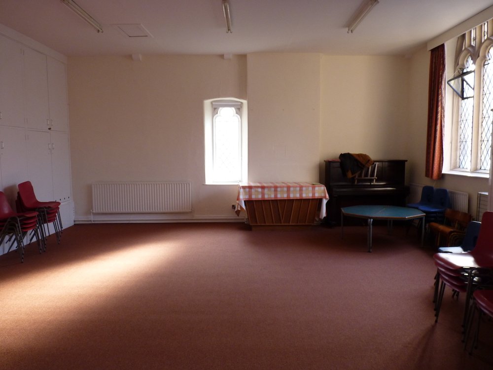 An empty room with a brown carpet, beige walls, and several chairs stacked and arranged around the edges. There are windows with stained glass and red curtains, a small table with a checkered tablecloth, and a piano with a chair on top.