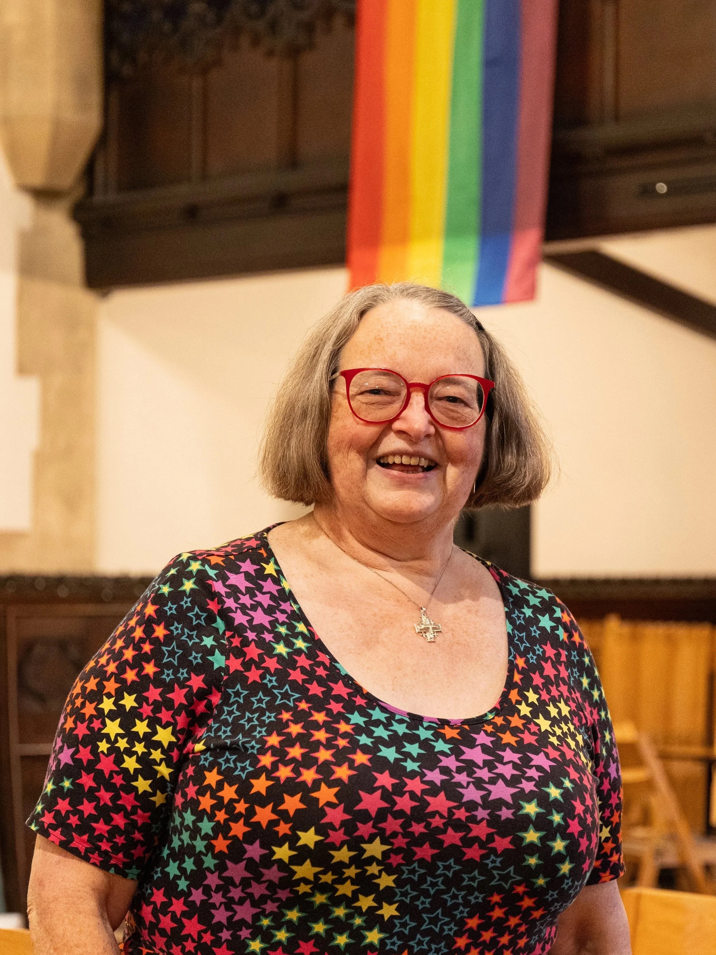 A middle-aged woman with short gray hair, wearing red glasses, and a colorful star-patterned dress, smiling at the camera. Behind her is a rainbow Pride flag hanging from the ceiling.