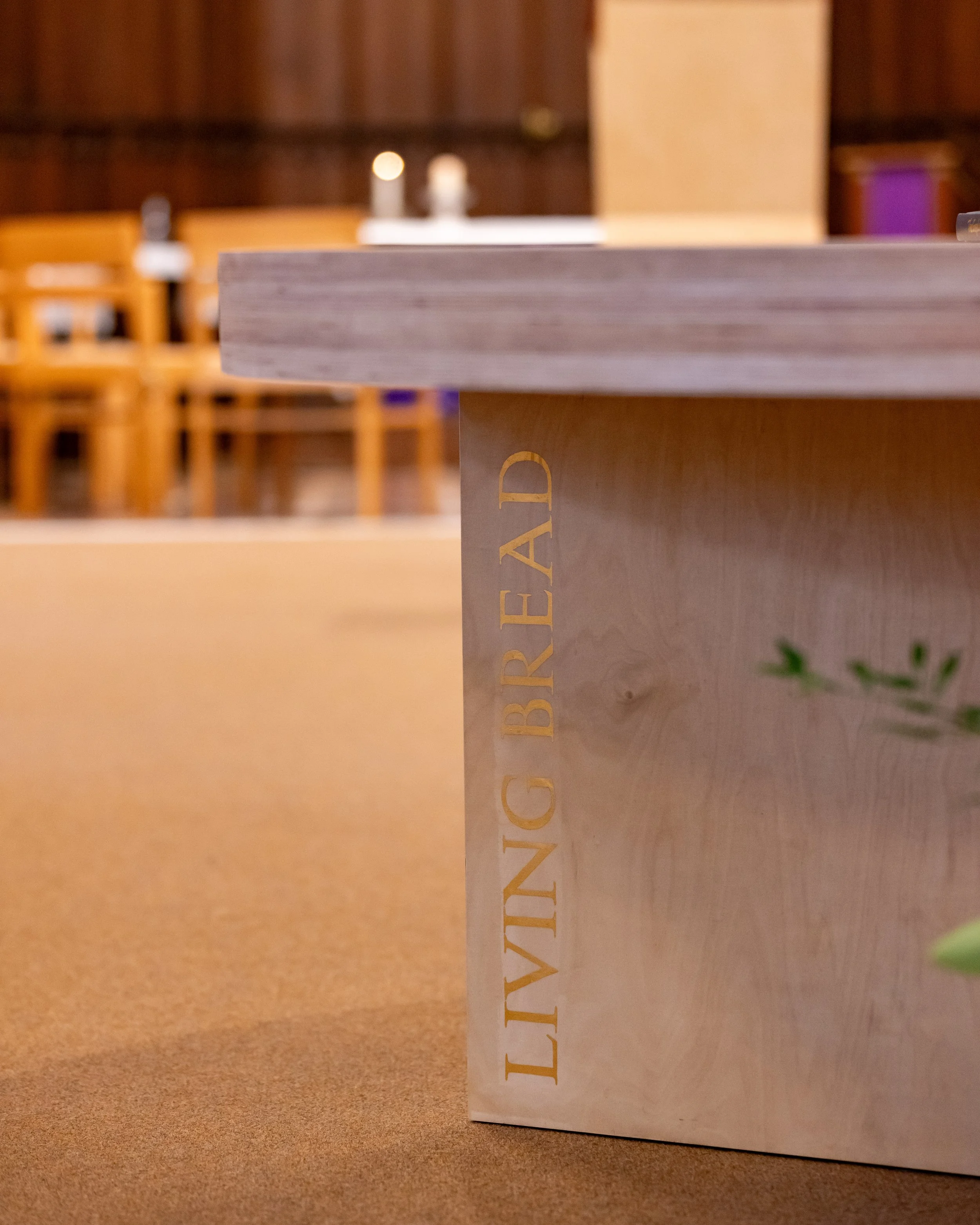 Close-up of a wooden table with gold embossed text that reads 'LIVING BREAD' on the side, with background chairs and tables in a blurred, warmly lit interior.