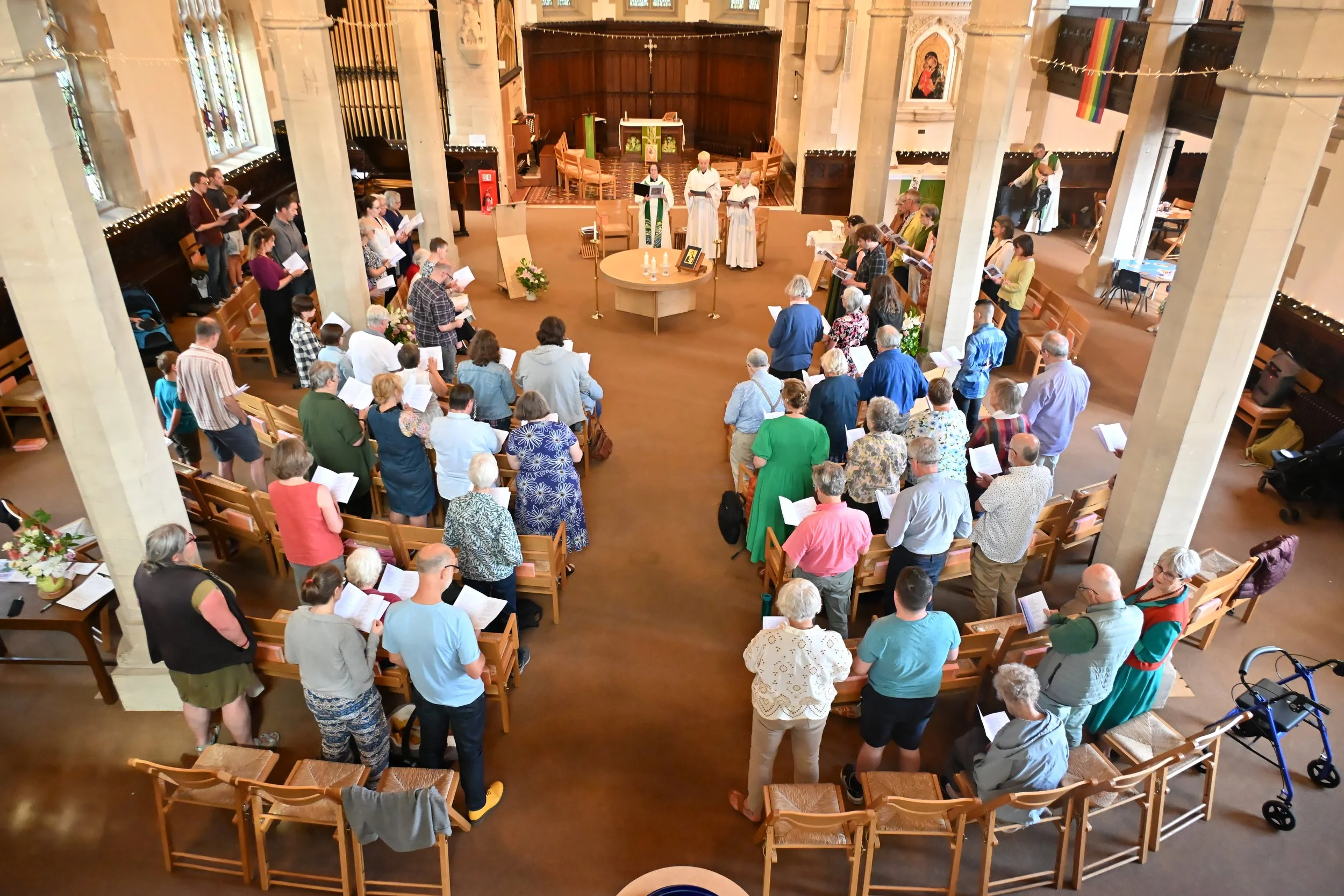 A congregation gathered inside a church for a service, with people standing and reading from books or hymns, facing the altar. The church interior is decorated with flowers and religious imagery, with stained glass windows on the sides.