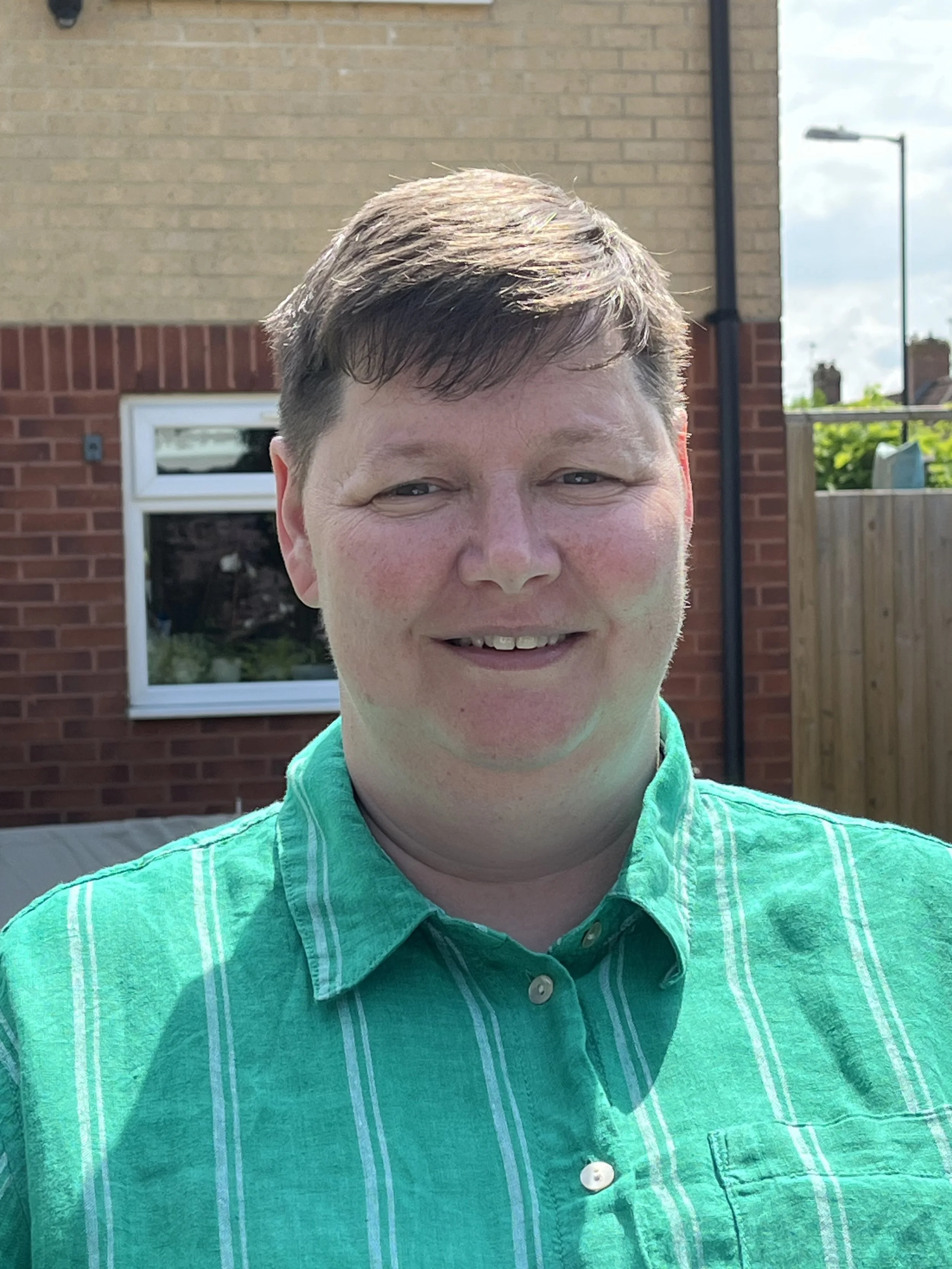A man with short brown hair and a fair complexion standing outdoors in front of a brick house, wearing a green vertically striped shirt and smiling.