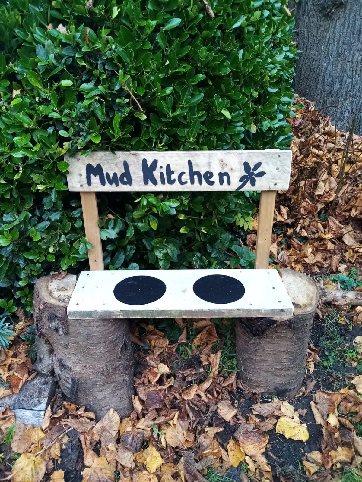 A small chair made from tree stumps and wood, with a sign reading 'Mud Kitchen' mounted on the backrest, placed outdoors among leaves and shrubbery.