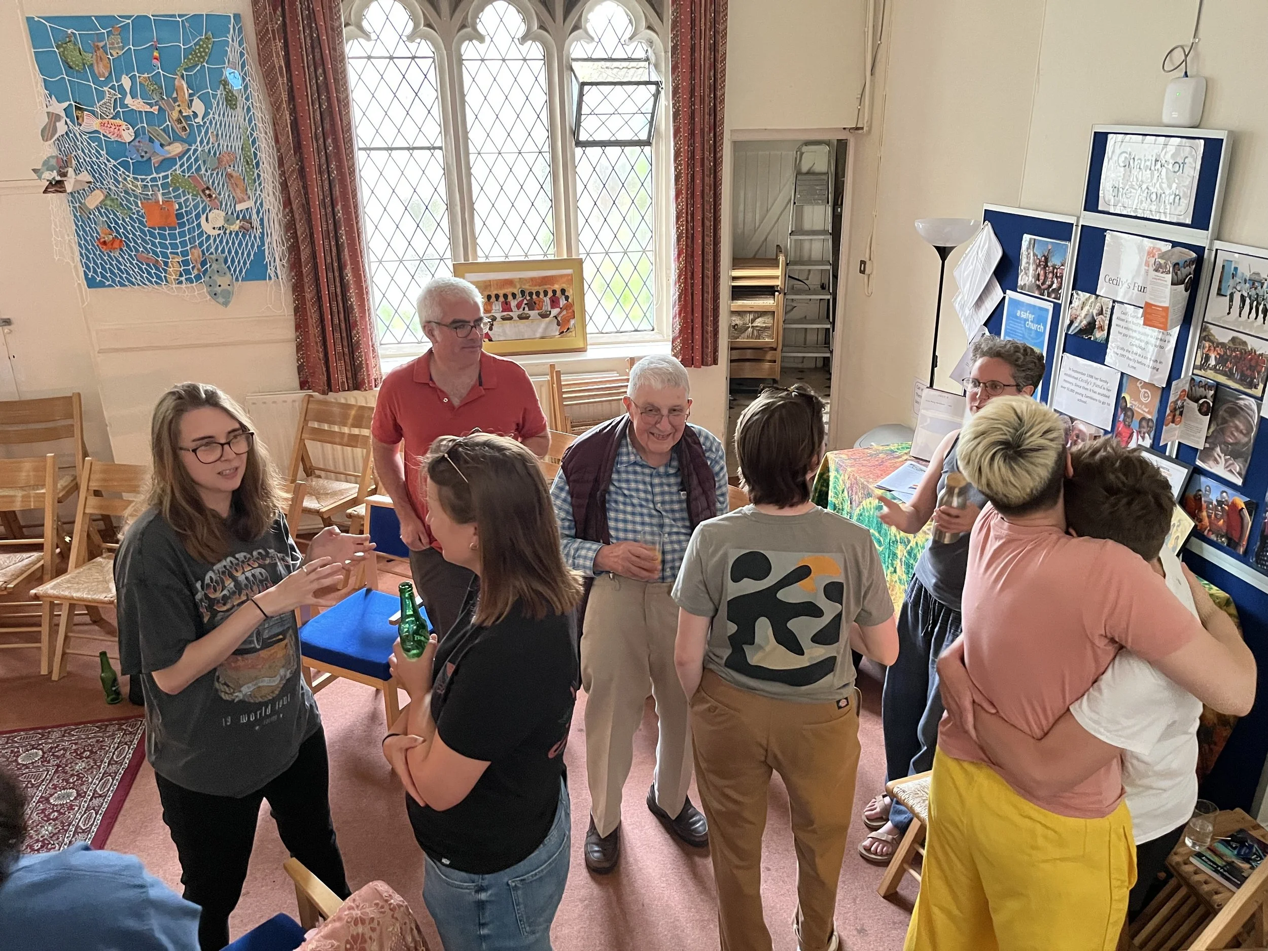 A group of people socializing and hugging indoors, with some holding drinks, in a room decorated with informational posters, photographs, and a colorful cloth on a table.