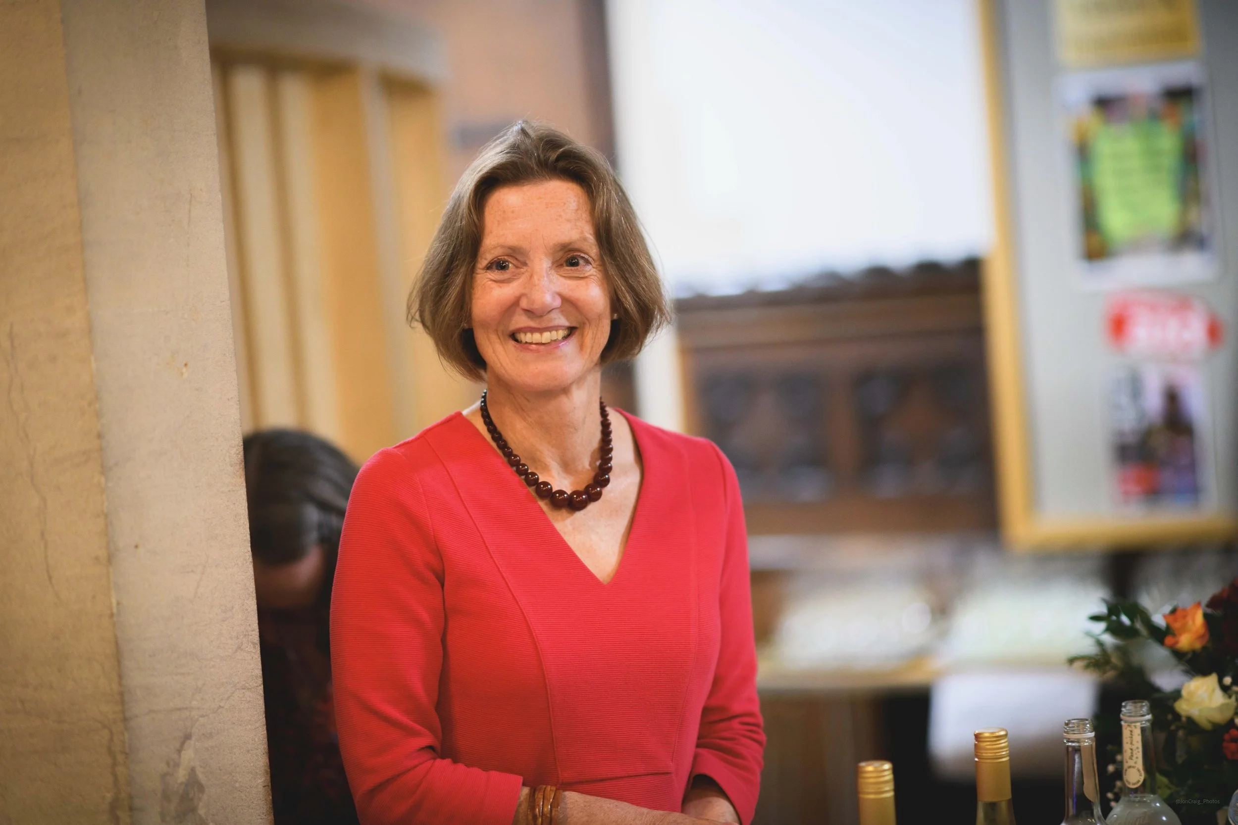 A woman with short brown hair, wearing a red dress and a dark beaded necklace, smiling in an indoor setting, with a blurred background that includes a cabinet and some bottles.