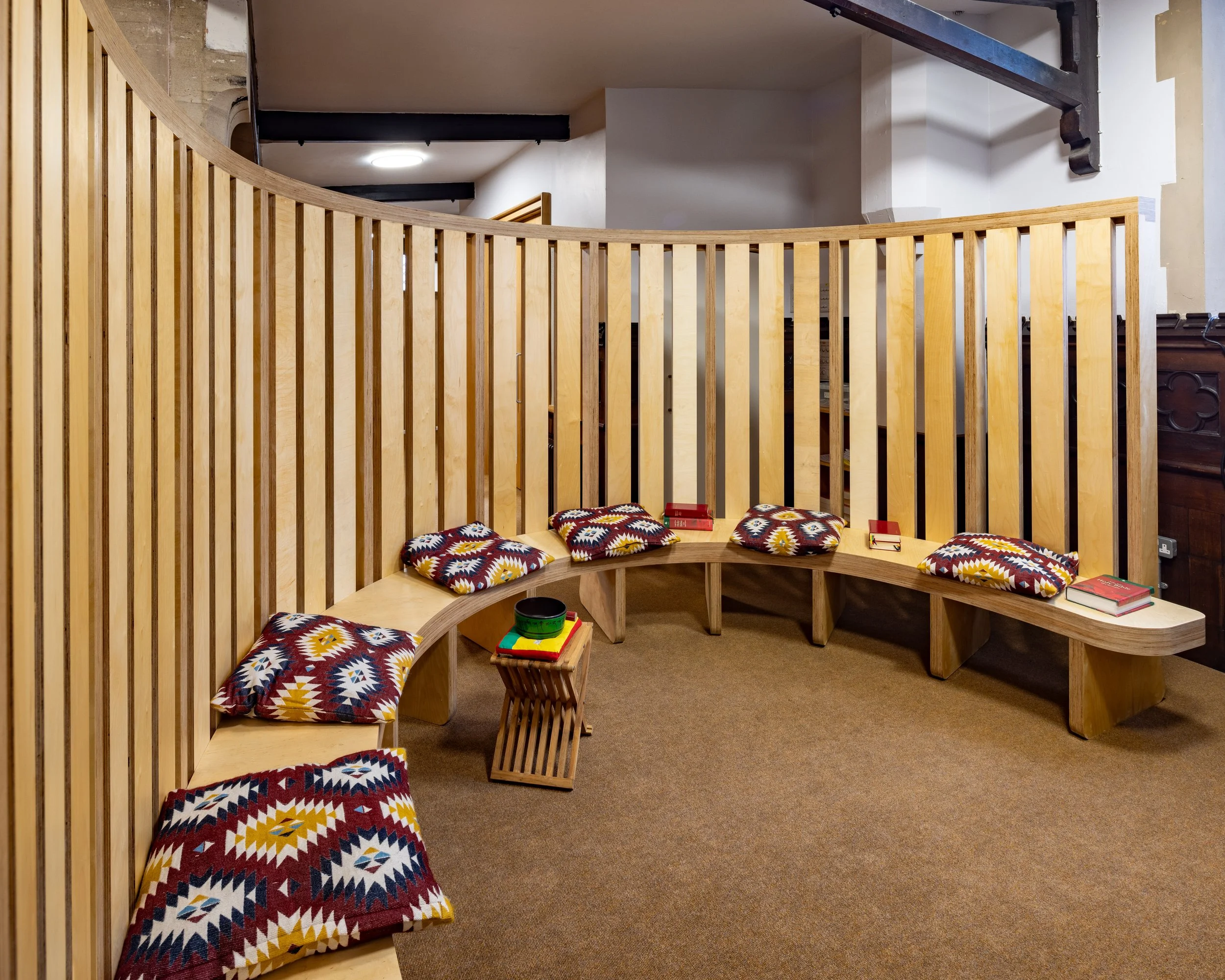 Indoor seating area with wooden curved bench, colorful patterned pillows, small table, and books on a brown carpet floor against a wooden slat wall and white ceiling.