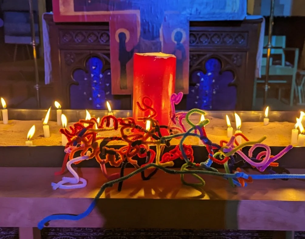 A table with a red candle, surrounded by colorful pipe cleaners shaped into various designs, with small lit candles around it. In the background, a wooden altar or furniture with religious or artistic symbols is visible.