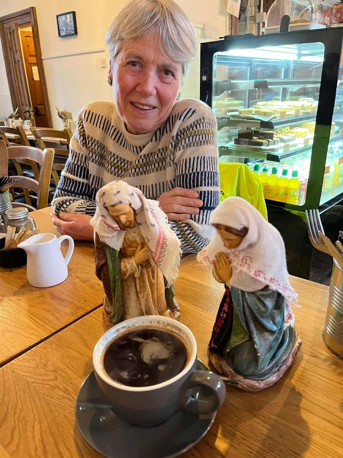An elderly woman sitting at a wooden table in a cafe, smiling at the camera, with two religious figurines of Jesus and Mary on the table, and a cup of coffee in the foreground. There is a glass display fridge in the background with various desserts inside.