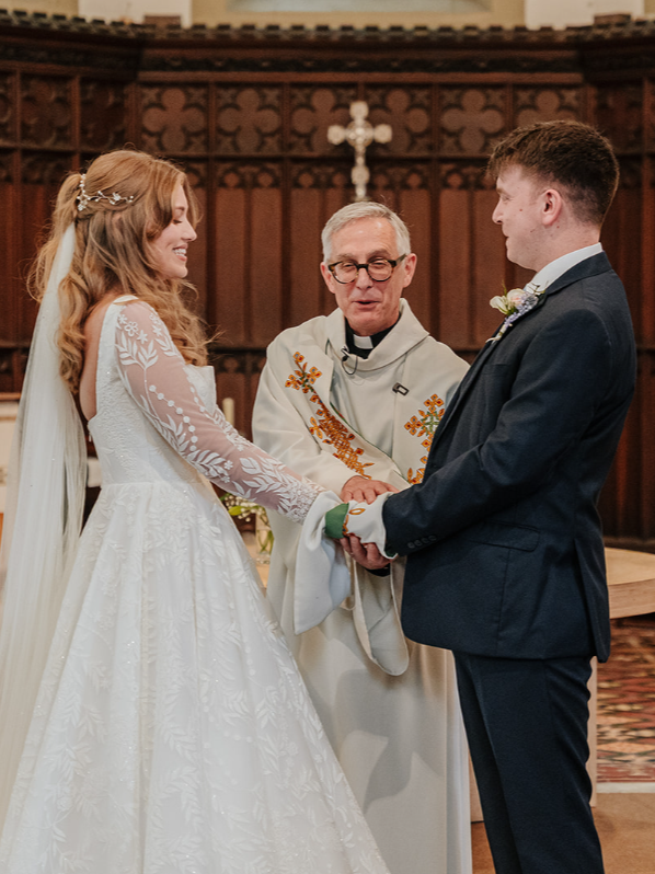 A bride and groom holding hands and smiling at each other during their wedding ceremony inside a church, with a priest officiating.