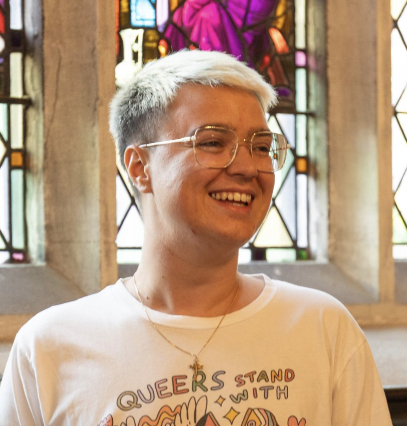 Person with short platinum blonde hair wearing glasses and a white t-shirt with colorful text, smiling inside a building with stained glass windows.