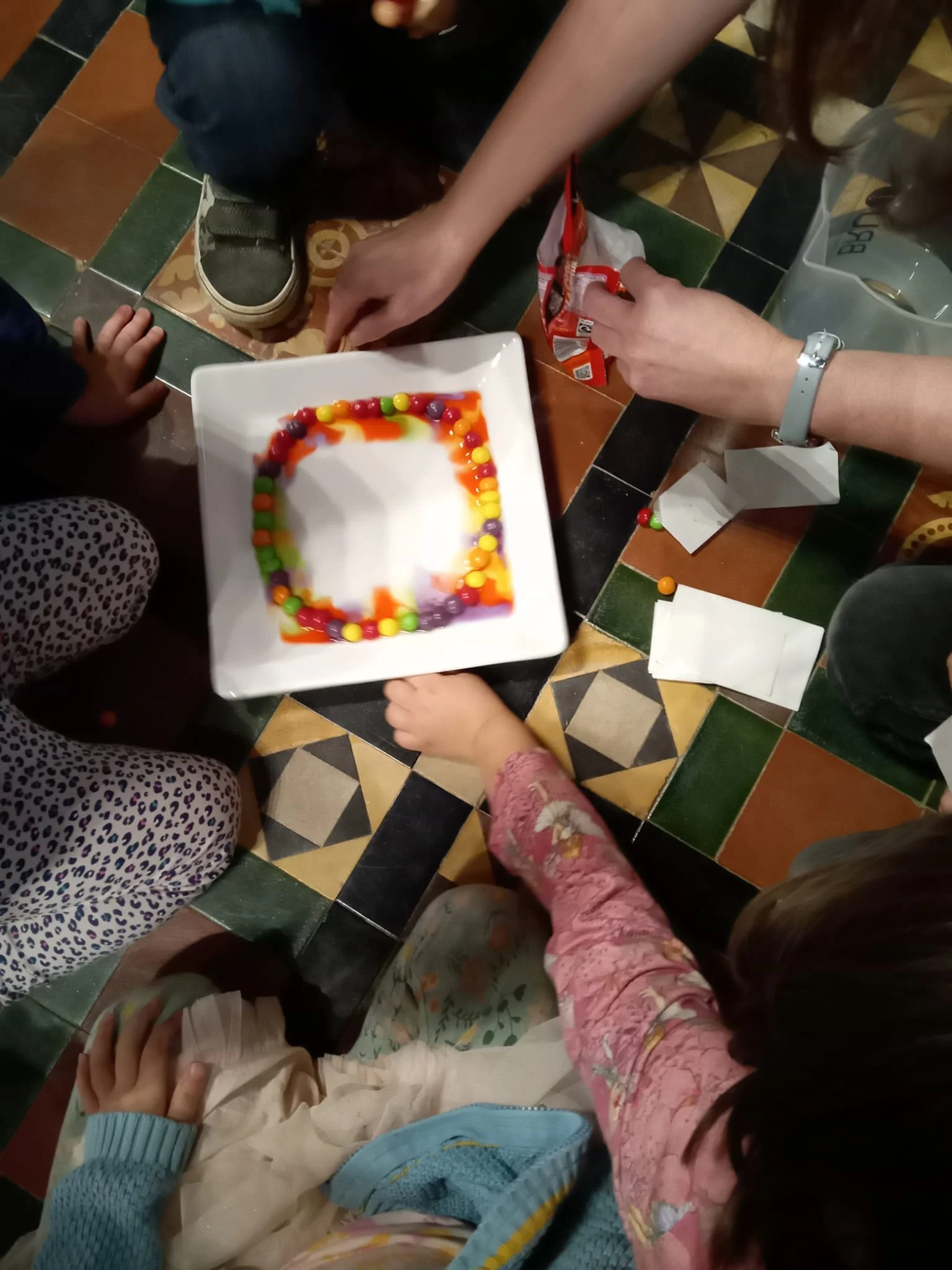 Children gathered around a table with a square white dish decorated with colorful candies and syrup.