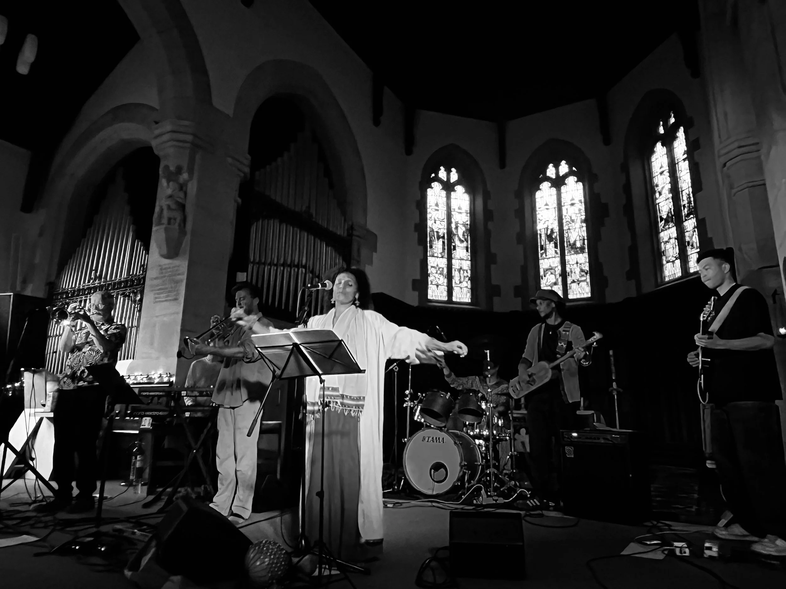 A band performing on stage inside a church, with stained glass windows in the background, and musicians playing various instruments including violin, guitar, and drums.