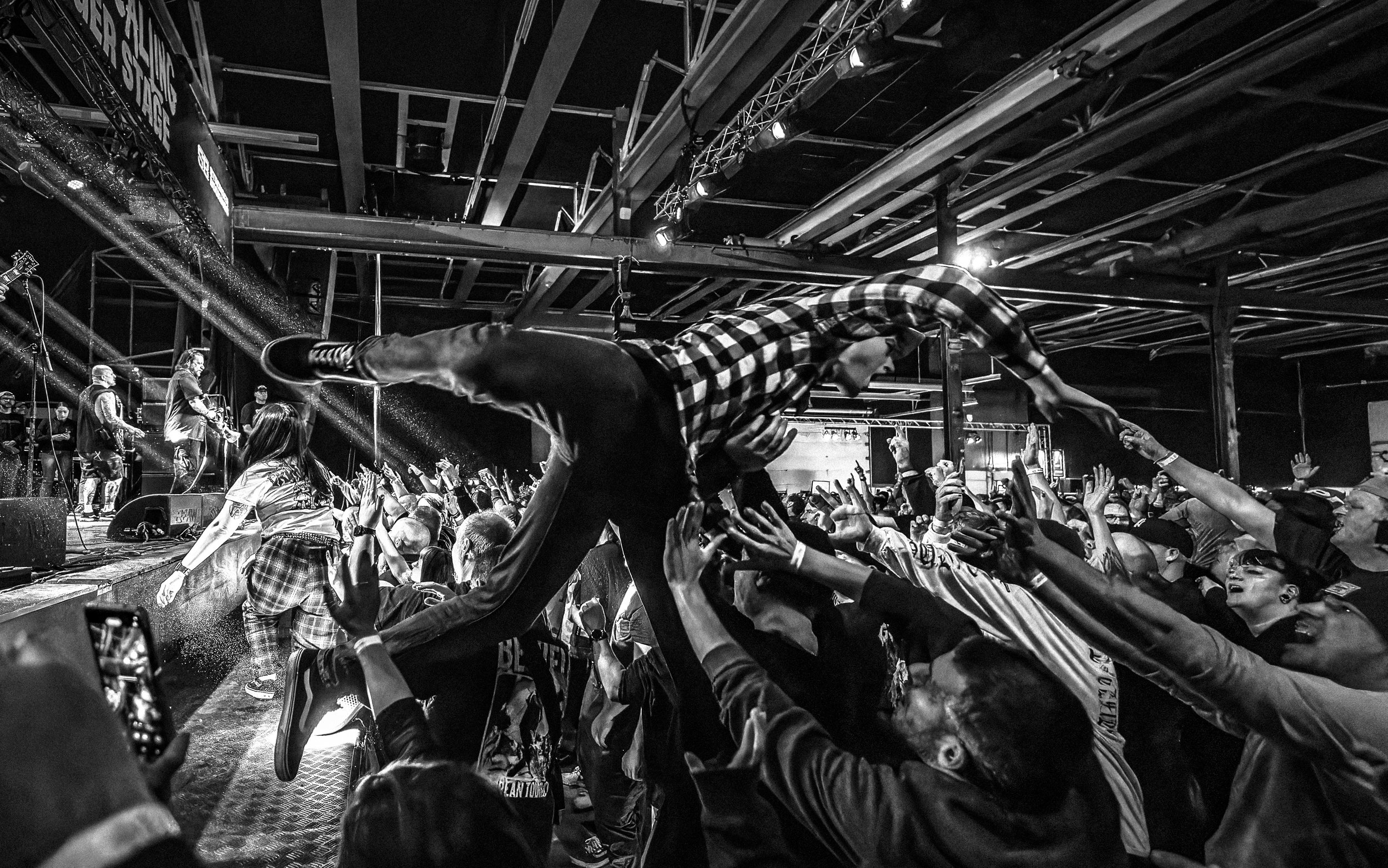 A concert scene in black and white with people reaching out to crowd surf as a performer crowd surfs over them. The audience is energetic, and the stage is visible with band members performing in the background.