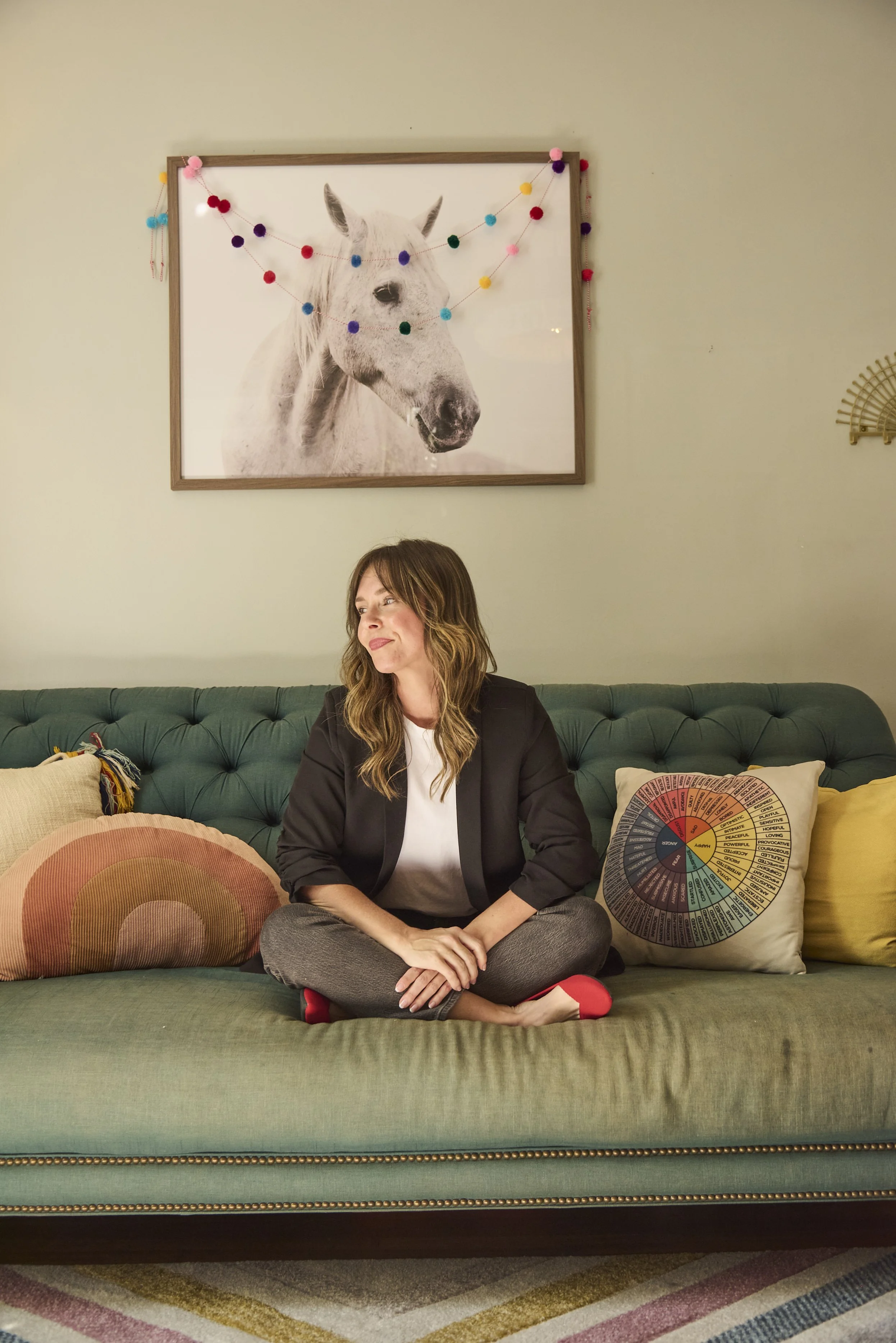 Woman sitting cross-legged on a green couch, surrounded by colorful pillows, in a room with a framed horse photograph on the wall decorated with a string of multicolored pom-pom garland.