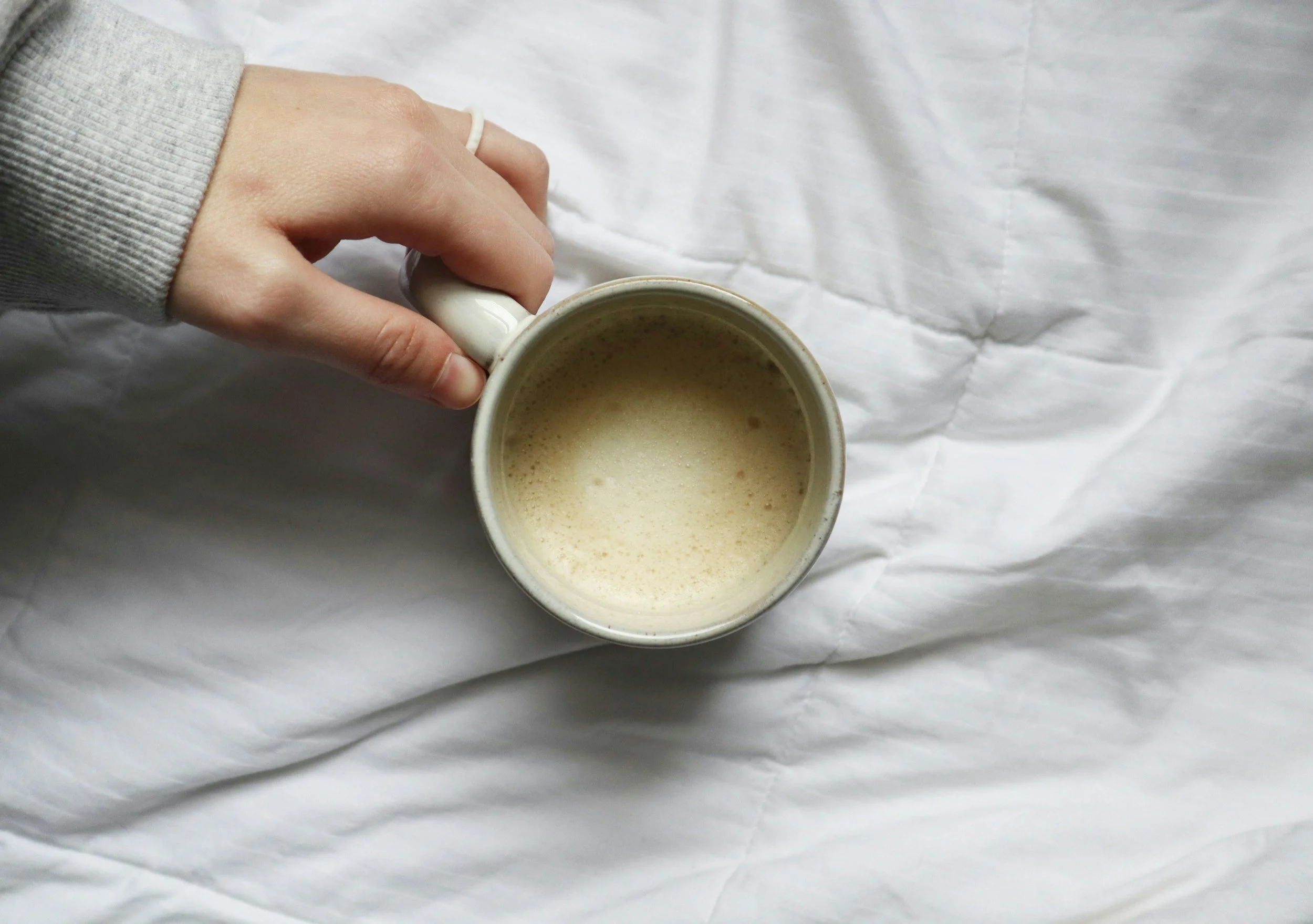 Overhead view of a hand holding a white ceramic mug filled with a frothy beverage, resting on a wrinkled white fabric surface.
