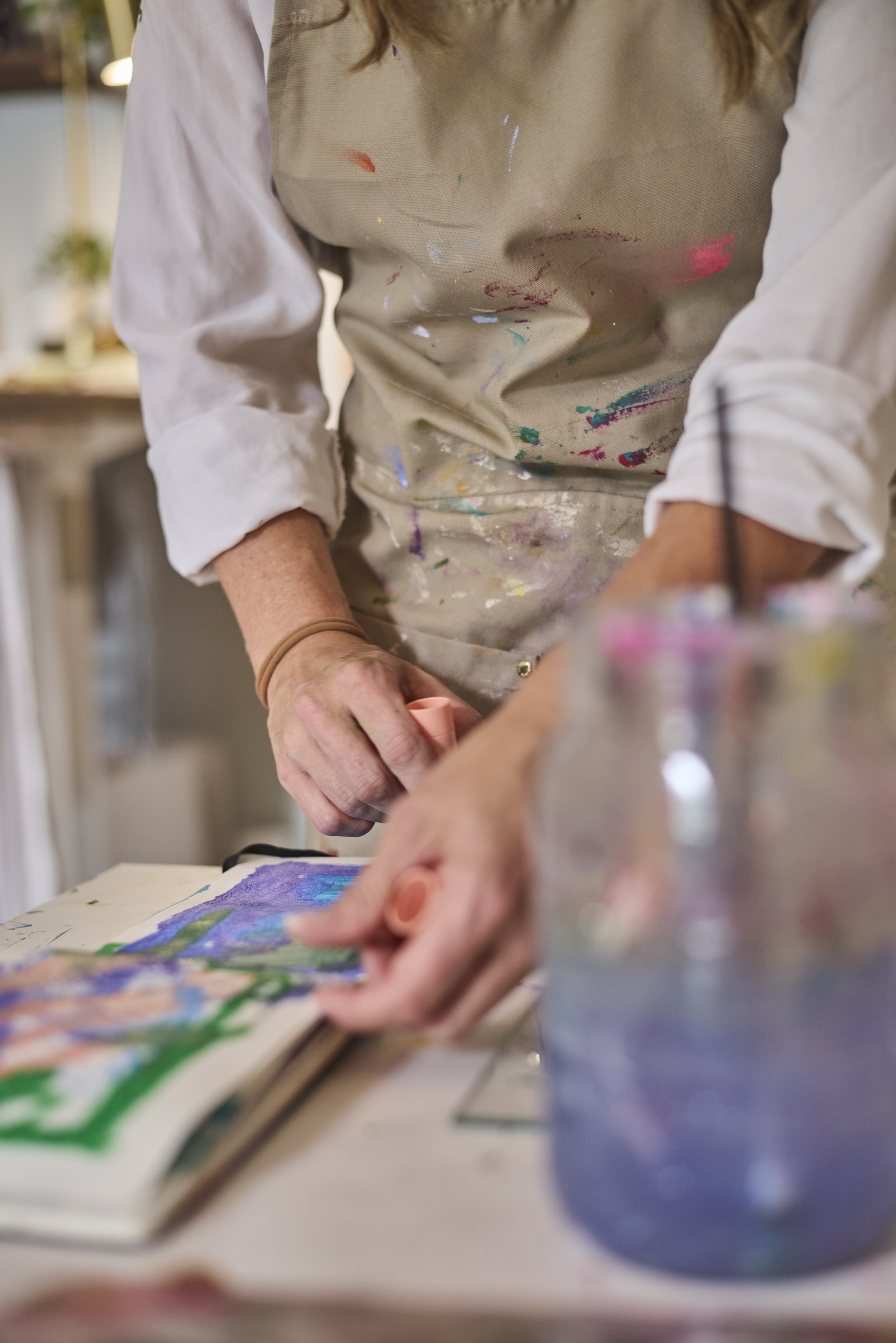 Person wearing a paint-splattered apron working on an art project, holding a small tool, with a colorful drawing or painting on the table.