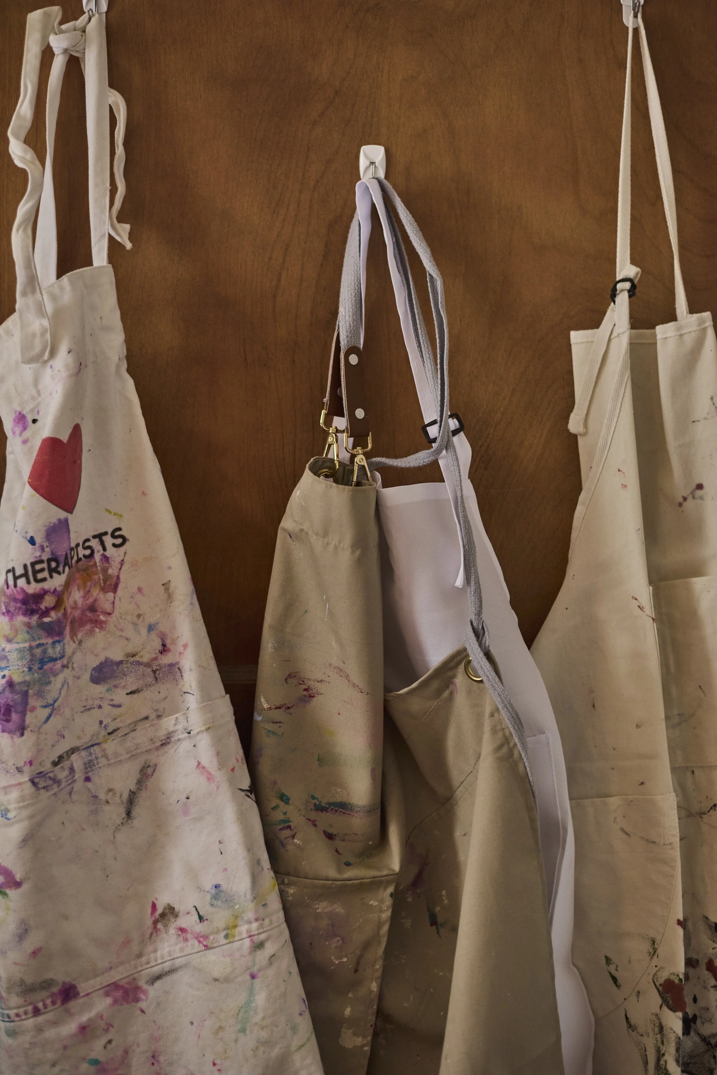 Paint-splattered aprons hanging on hooks on a wooden wall, with various art supplies attached or hanging.