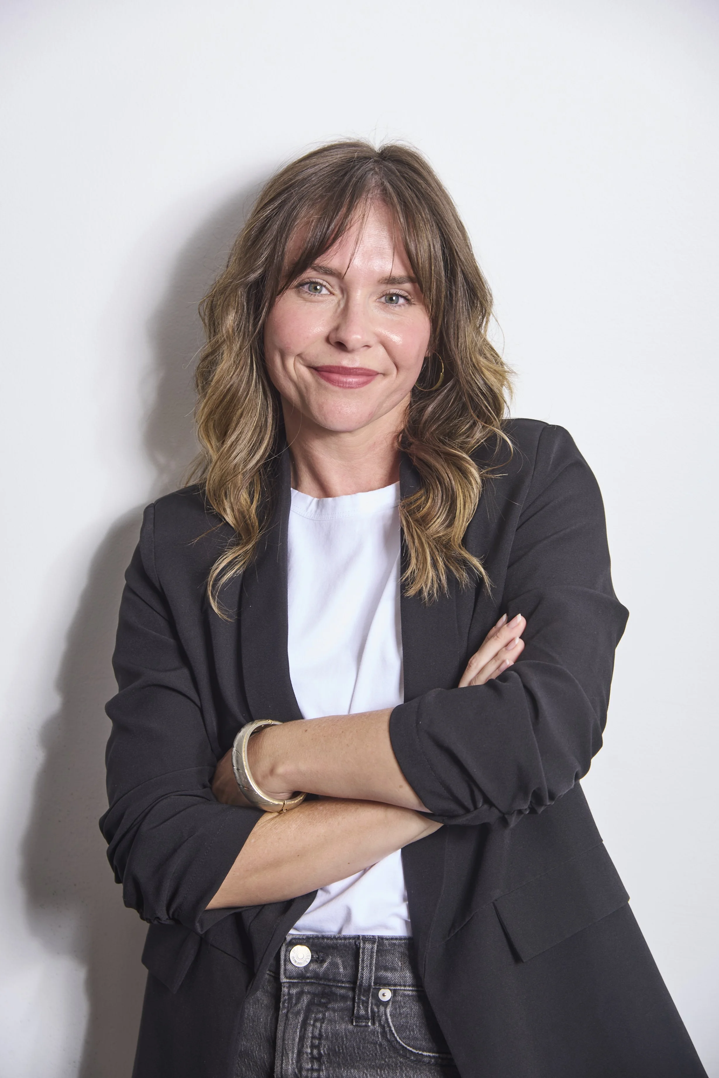A woman with shoulder-length wavy brown hair, wearing a black blazer over a white shirt, standing with arms crossed against a plain white wall.