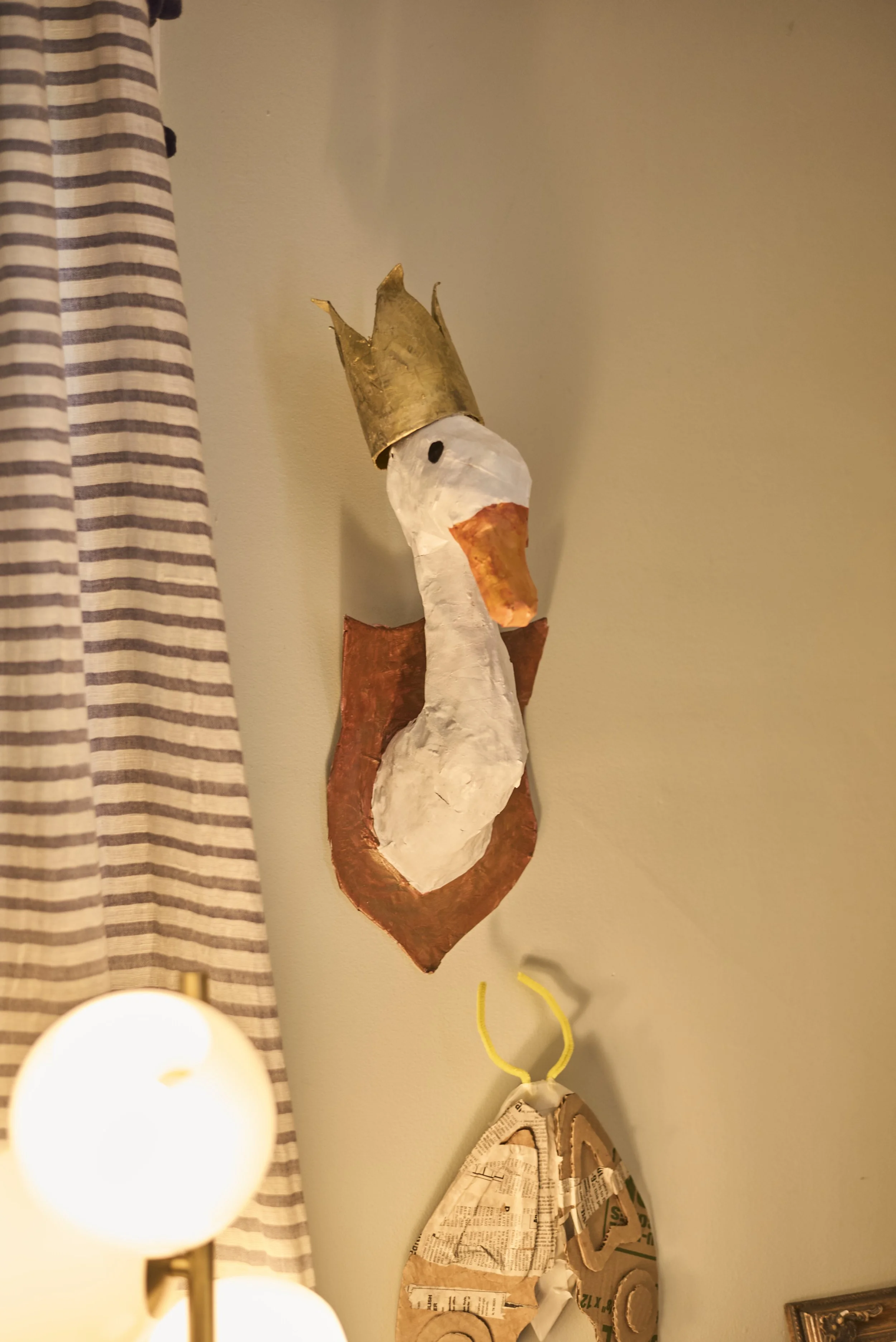 A handmade decorative wall hanging of a stork wearing a golden crown, with white and orange features, mounted on a wall next to a striped curtain and a paper mache butterfly.
