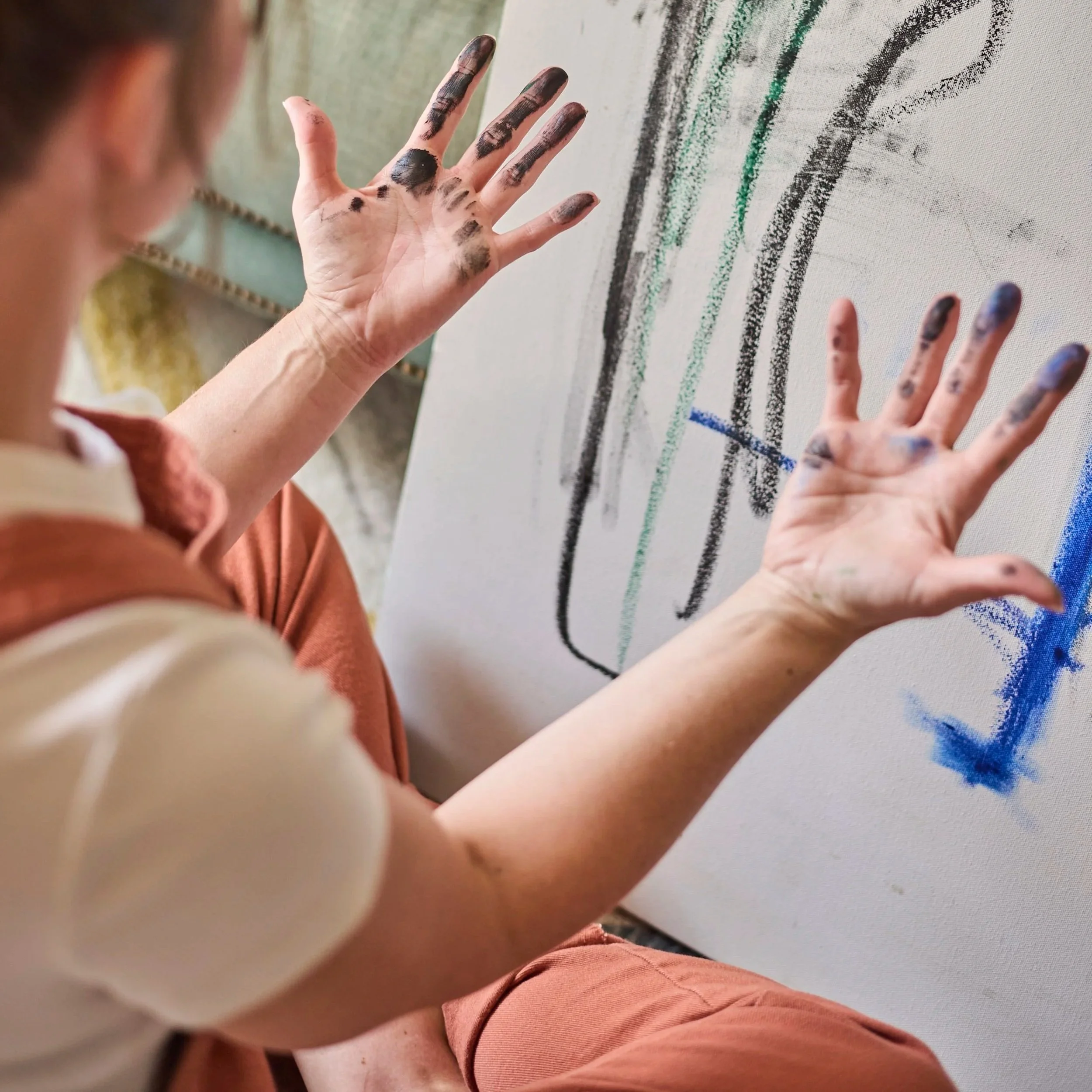 A person with paint on their hands, sitting in front of an abstract painting with colorful brushstrokes.