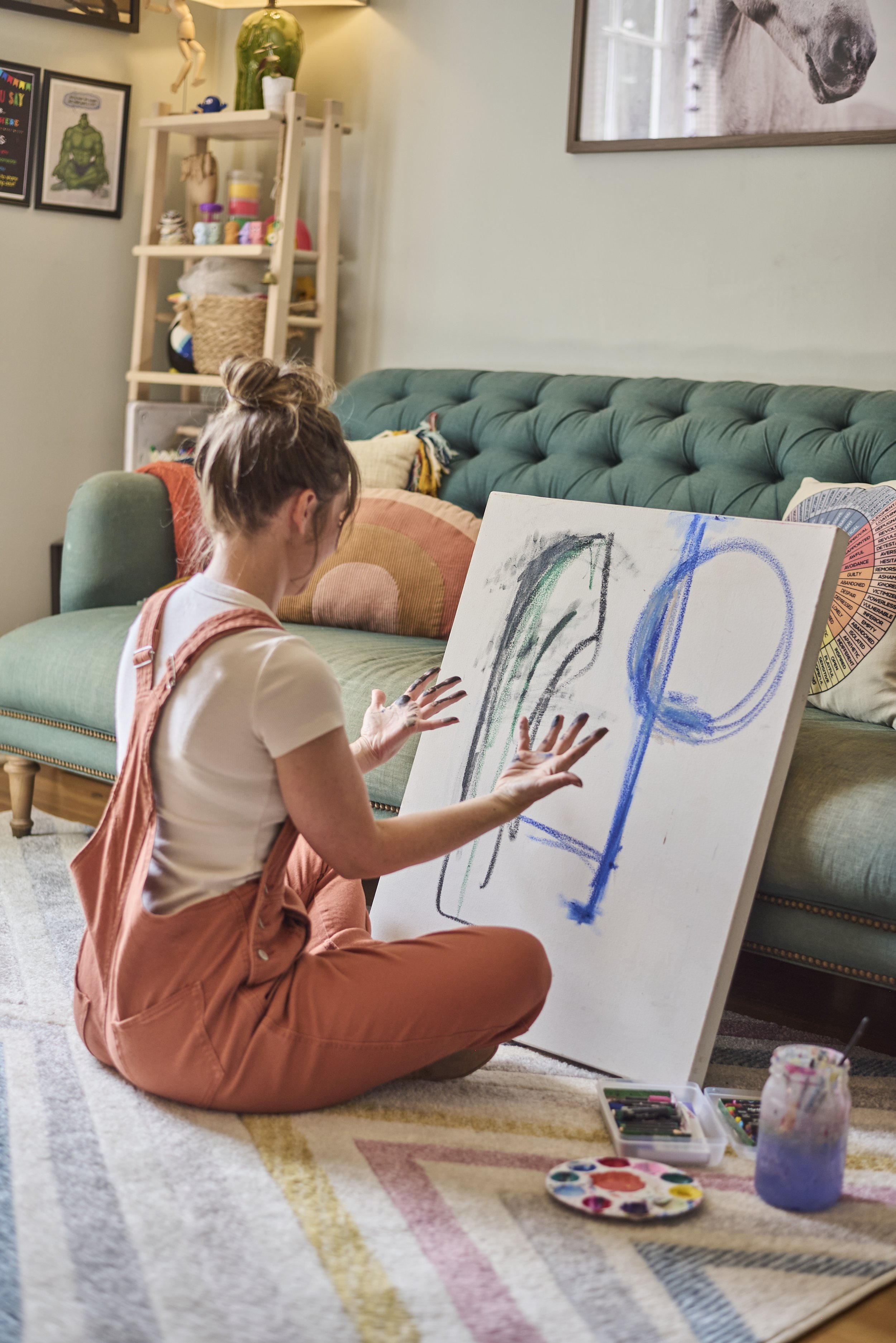 A woman sitting on the floor in front of a large white canvas, painting with colorful pastels or chalk. She has her hair in a bun, is wearing a white shirt and pink overalls, and is surrounded by art supplies including a paint palette, colored pastels, and jars of water. The room features a green sofa, decorative pillows, a wooden shelf with plants, art prints on the wall, and a patterned rug.