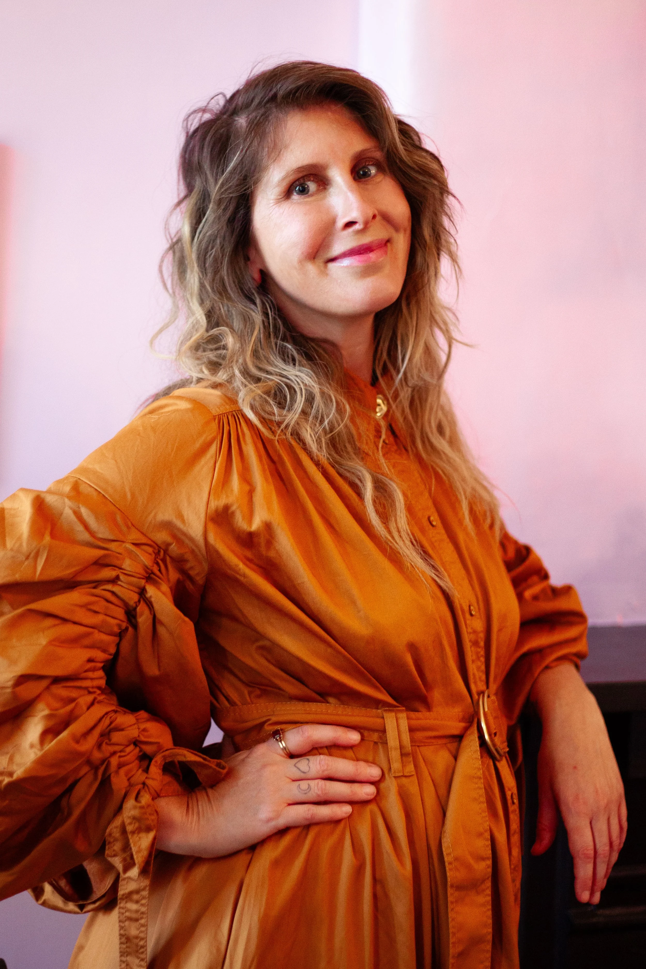 Sammy-Jo, with shoulder-length wavy hair, wearing an orange dress with long sleeves, smiling and posing with her hand on her hip against a pinkish background.