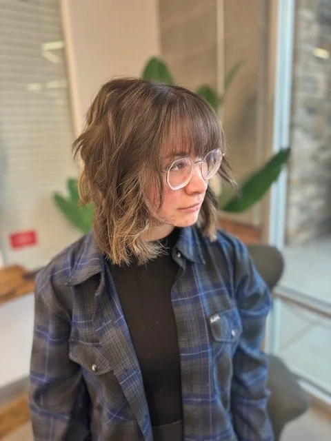 A woman with shoulder-length wavy hair and glasses, wearing a black shirt and blue plaid shirt, is looking to her left indoors near a window with a large plant in the background.