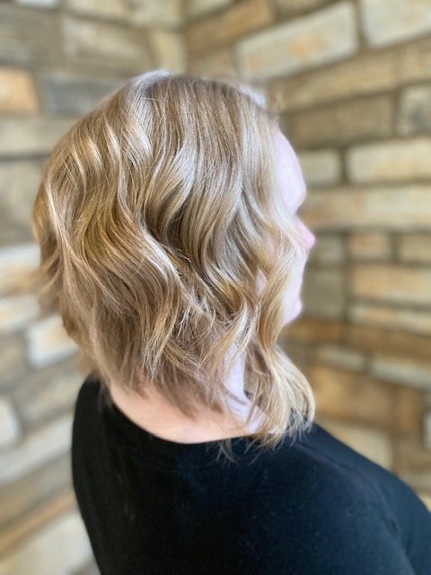 Side view of a woman with shoulder-length, wavy blonde hair, standing in front of a brick wall.