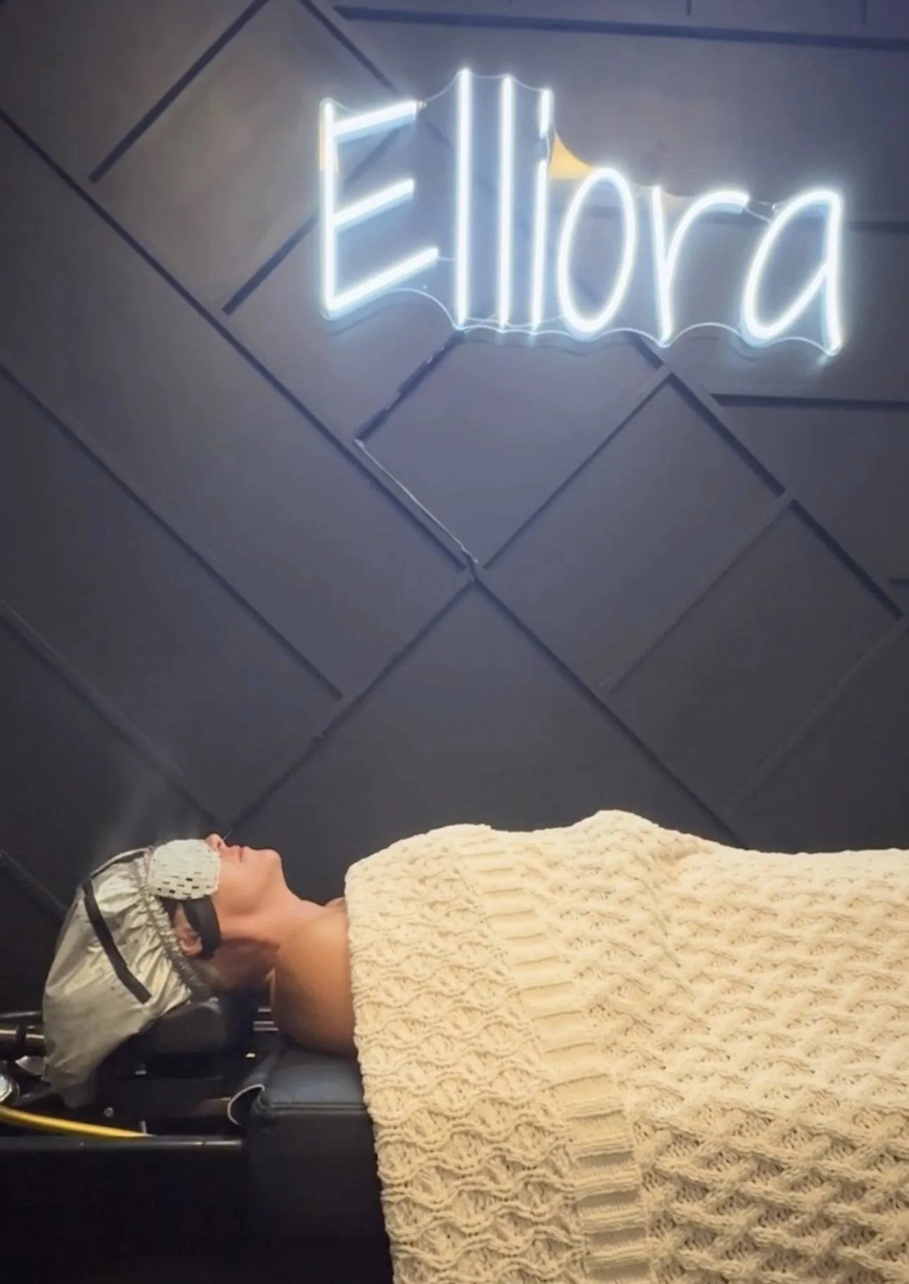 A woman lying down under a blanket, receiving a spinal treatment in a clinic, with a neon sign above that reads 'Elliora'.