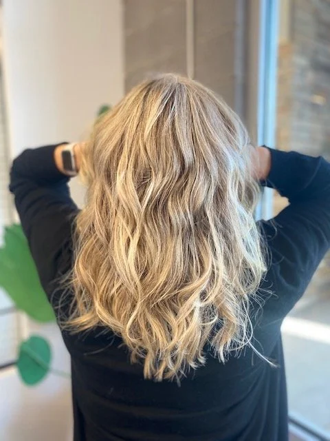 Back view of a woman with shoulder-length, wavy blonde hair, wearing a black top, inside a modern room with a plant and large window.