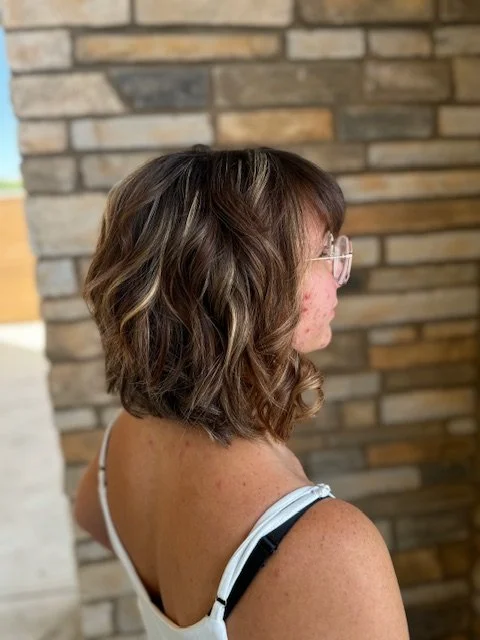 Side view of a woman with curly brown hair and glasses, wearing a white top, standing indoors in front of a brick wall.
