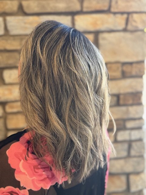 Side view of a woman with shoulder-length wavy blonde hair wearing a black top with a pink and red floral pattern, standing in front of a brick wall.