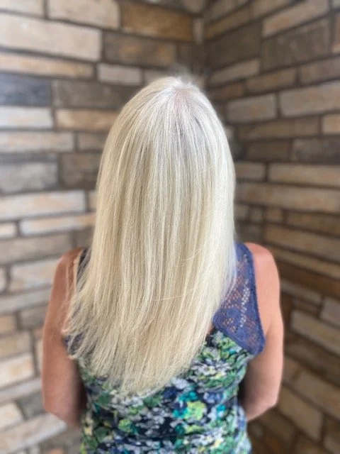 Back view of a woman with long, straight, blonde hair, wearing a floral top, in front of a brick wall.