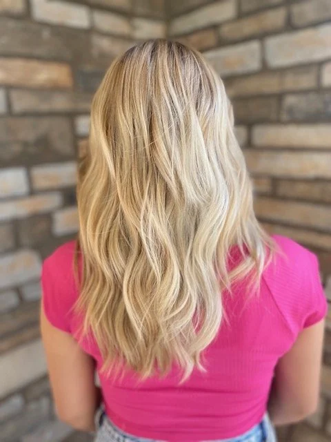 Back of a woman with long, wavy blonde hair wearing a pink top, standing in front of a brick wall.