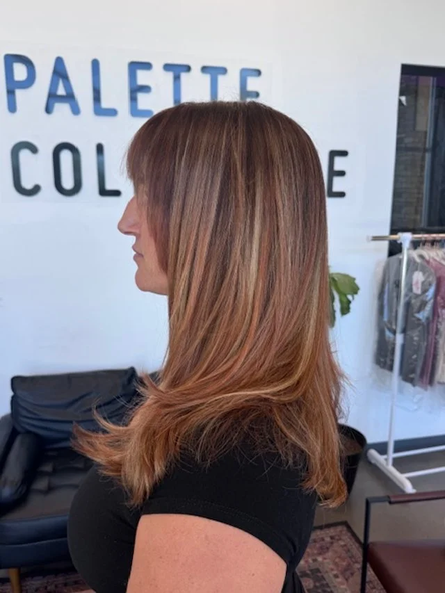 Side view of a woman with straight, shoulder-length, auburn hair in a salon or store with a white wall and clothing rack in the background.