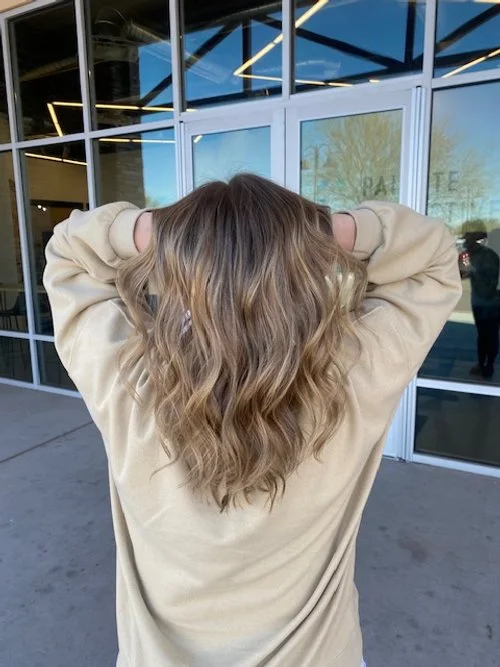 Person with wavy blonde hair standing outside a glass building, facing away from the camera with hands behind head.