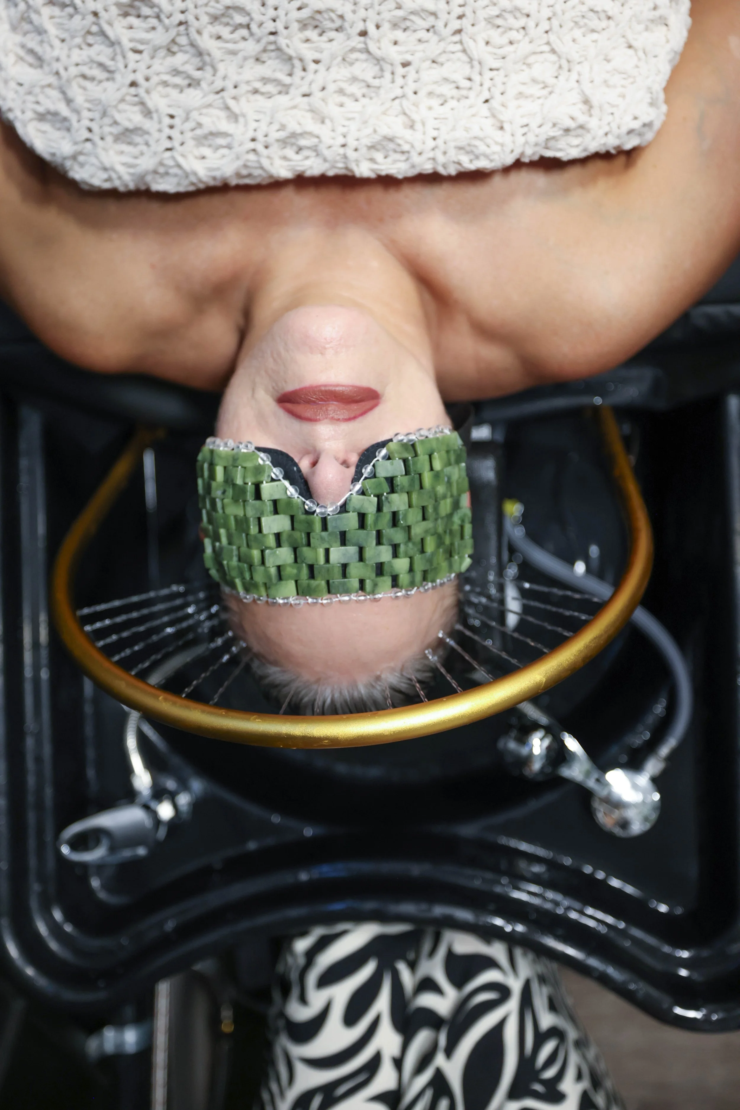 Close-up of a woman with a headscarf and makeup, lying in a specialized chair with a golden frame, wearing a green beaded mask covering her eyes, and surrounded by medical or spa equipment.