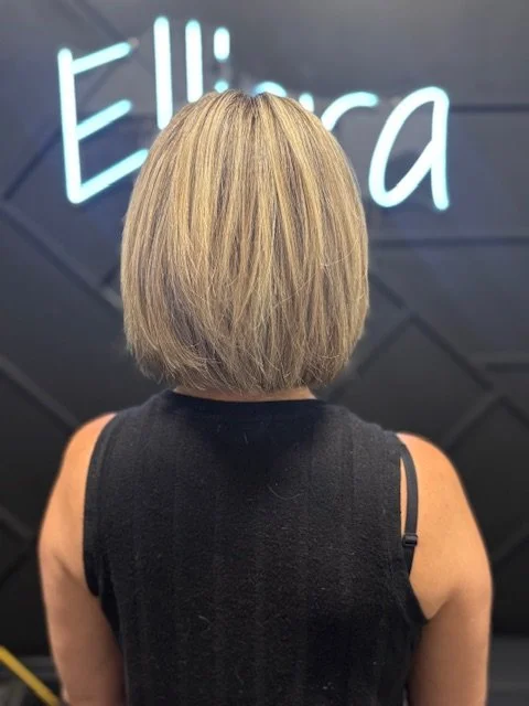 Back of a woman with blonde, layered bob hairstyle, wearing a black sleeveless top, standing in front of a dark wall with neon sign that reads 'Eliza'.