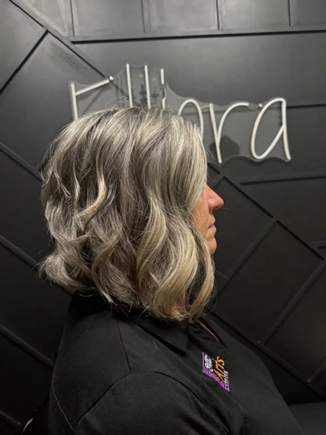 Profile of a woman with shoulder-length wavy gray hair, standing indoors in front of a black wall with white cursive lettering.