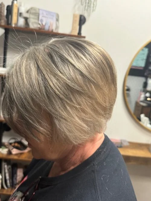 Side view of a woman with short blonde hair in a salon with a mirror and shelves in the background.