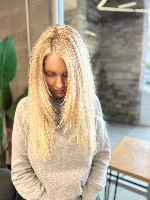 A woman with straight blonde hair looking down, wearing a gray sweatshirt, standing indoors near a window and a large green plant.