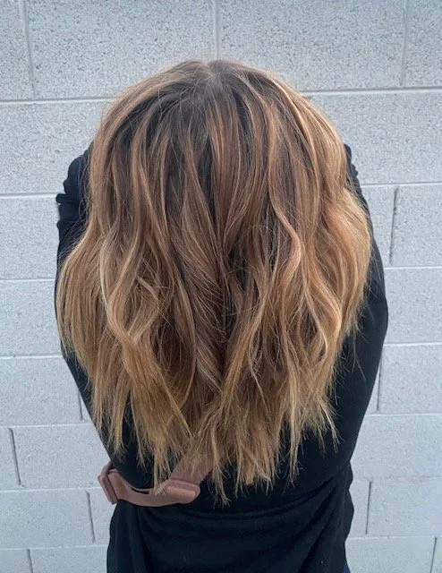 Person with long, wavy, light brown hair leaning forward against a gray concrete block wall, wearing a black top.