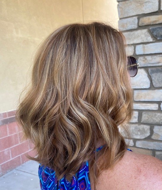 Side view of a woman with shoulder-length wavy blonde hair, wearing sunglasses and a blue patterned top, standing outdoors near a stone wall.