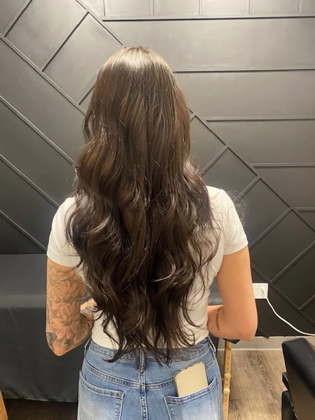 Woman with long wavy brown hair standing with her back to the camera in a room with black paneled walls, wearing a white t-shirt and blue jeans.