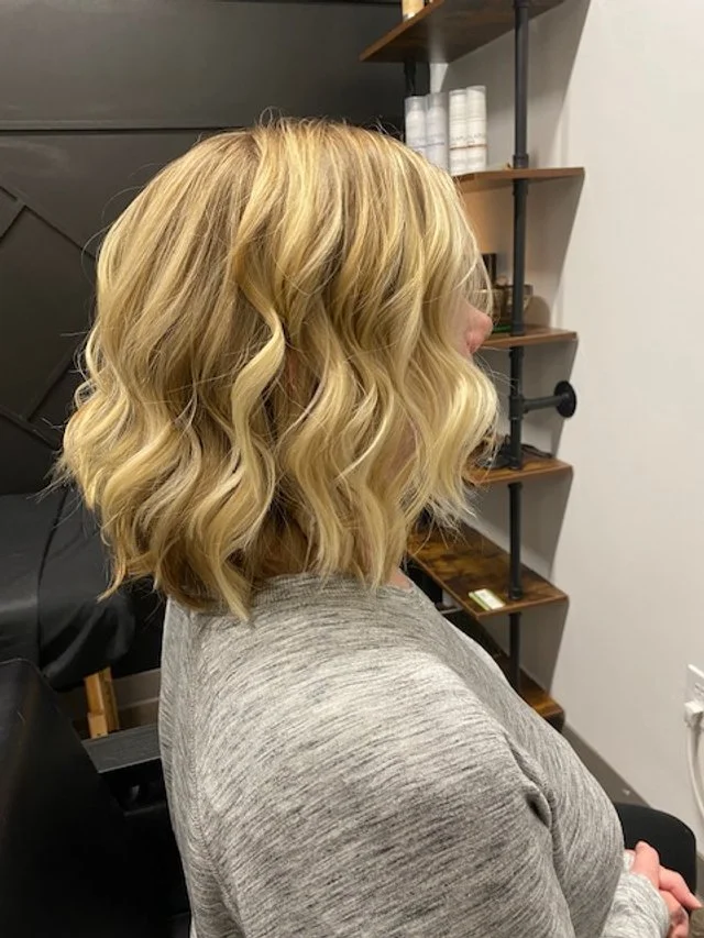 Side view of a woman with shoulder-length blonde wavy hair, sitting in a salon with shelves and styling products in the background.